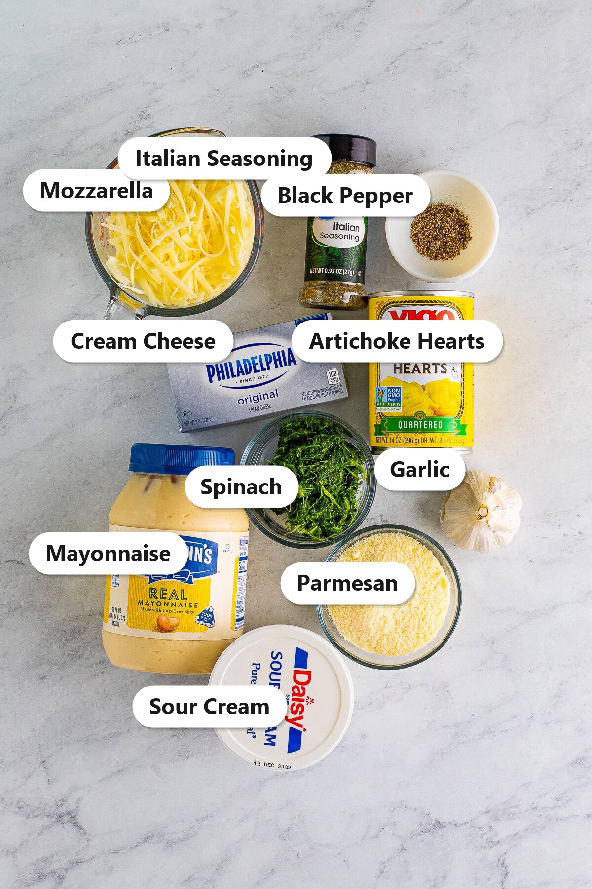 Labeled ingredients to make spinach artichoke dip including artichoke hearts, spinach, cream cheese, mozzarella, parmesan, and sour cream.
