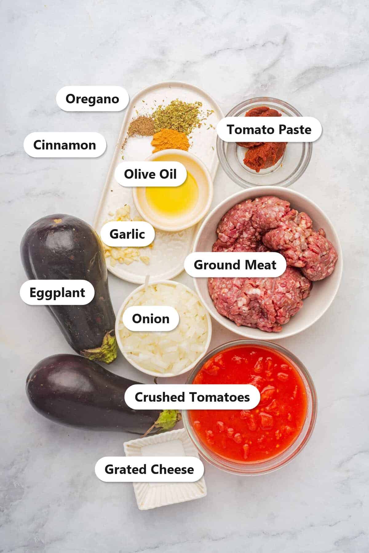 Labeled ingredients to make moussaka including eggplant, ground meat, crushed tomatoes, onion, garlic, and cheese.