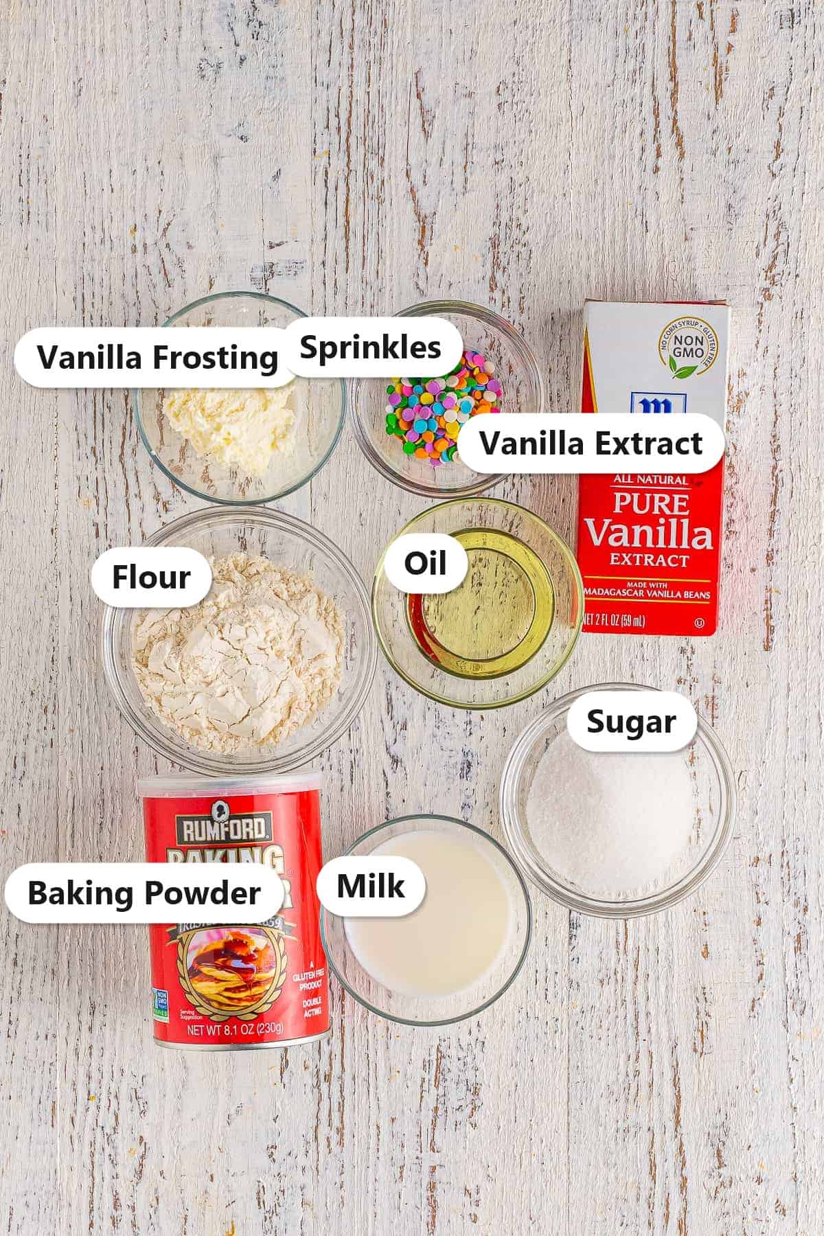 Labeled ingredients for vanilla mug cake including flour, sugar, baking powder, milk, oil, vanilla extract, frosting, and sprinkles.