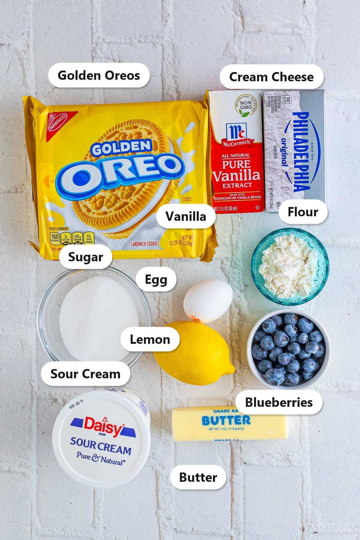 Labeled ingredients for lemon blueberry cheesecake bars including Golden Oreos, cream cheese, sour cream, sugar, eggs, flour, lemon, blueberries, vanilla, and butter.