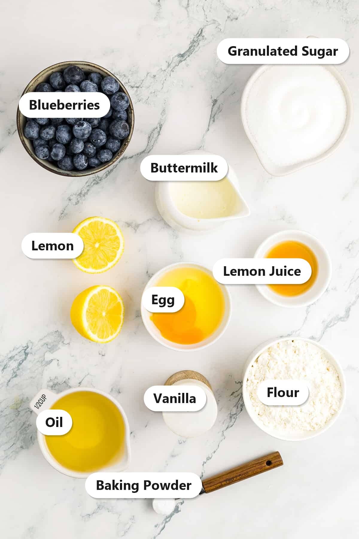 Ingredients to make lemon blueberry muffins labeled on a marble surface: blueberries, granulated sugar, buttermilk, lemon, egg, lemon juice, oil, vanilla, flour, and baking powder.