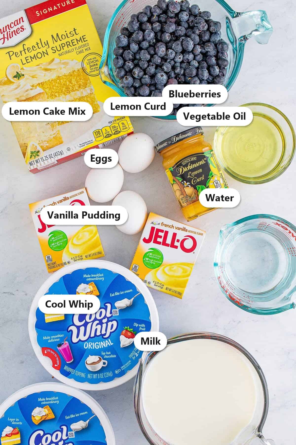 Labeled ingredients for lemon blueberry trifle including lemon cake mix, blueberries, vanilla pudding, lemon curd, Cool Whip, eggs, oil, and milk.