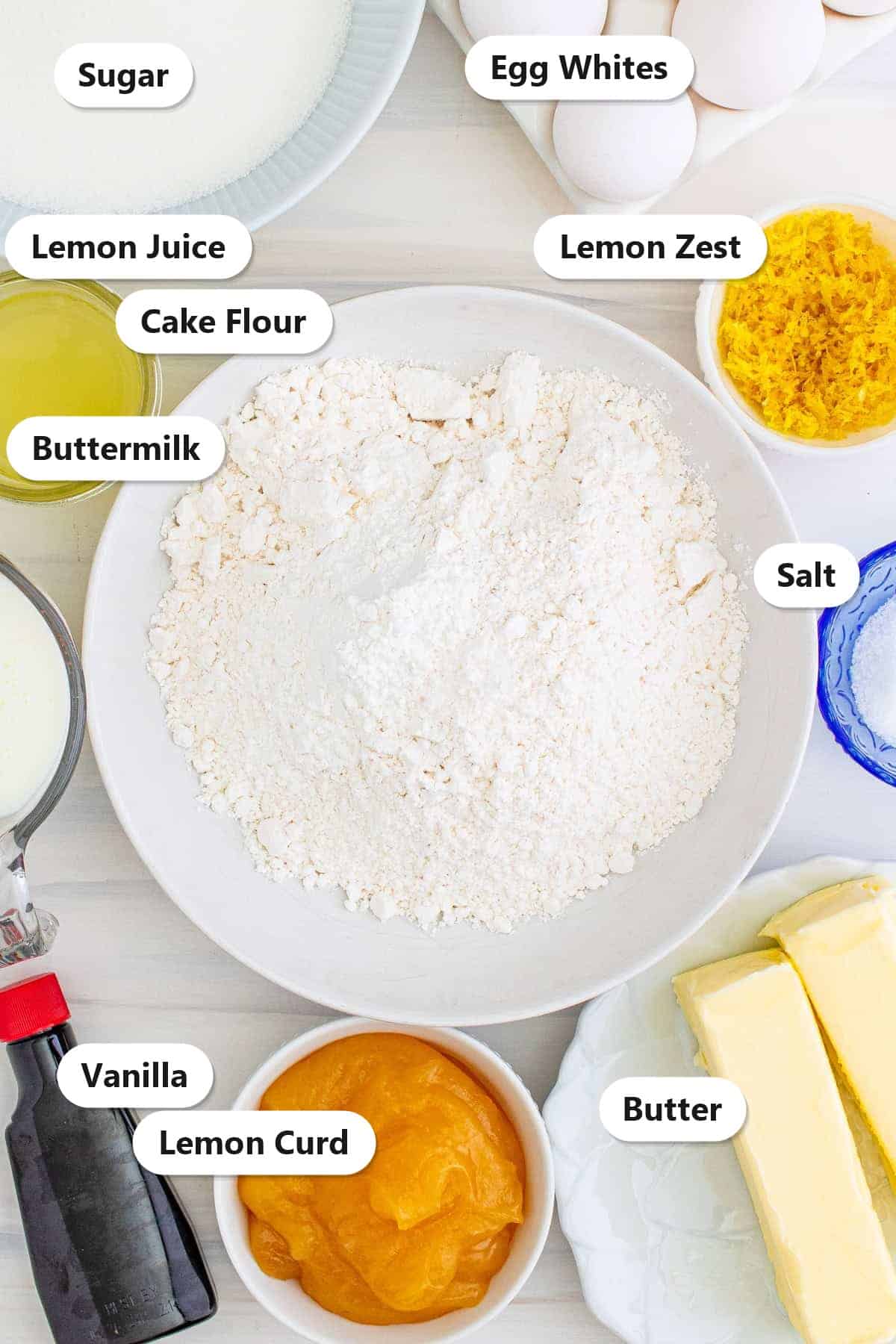 Labeled ingredients to make lemon cake including cake flour, sugar, egg whites, buttermilk, lemon juice, lemon zest, lemon curd, and butter.