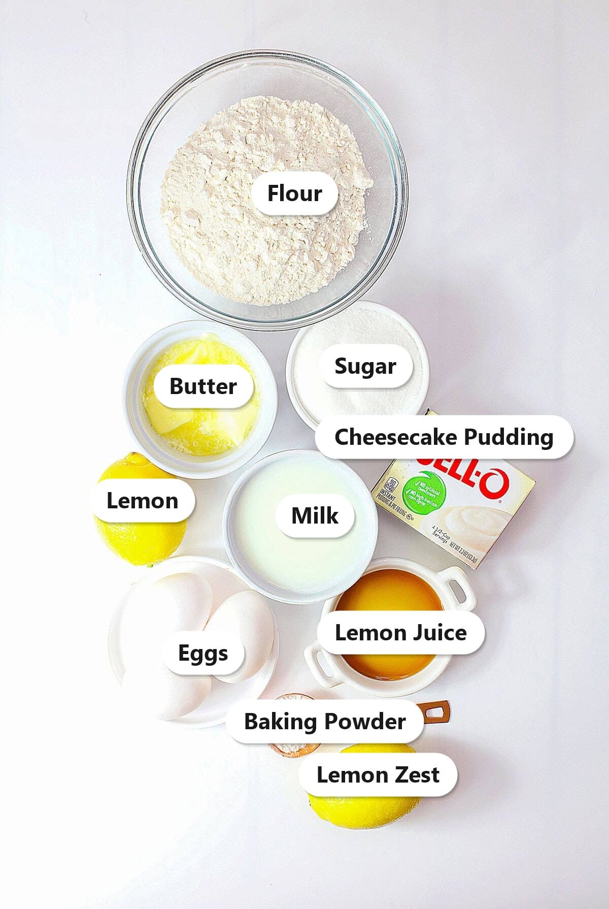 Labeled ingredients for lemon cheesecake cupcakes including flour, butter, sugar, cheesecake pudding, lemon, milk, eggs, lemon juice, baking powder, and lemon zest.