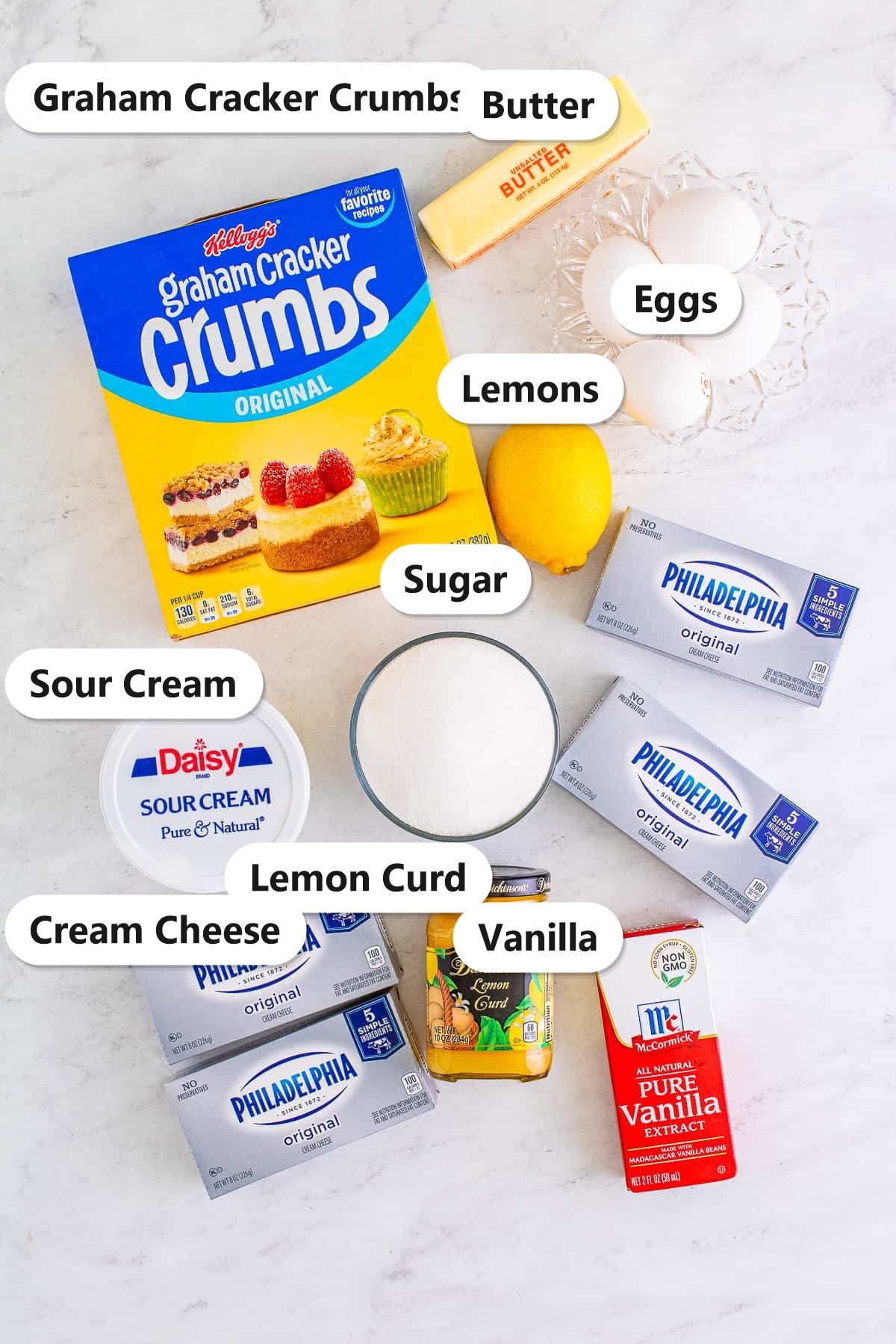 Lemon Cheesecake ingredients including graham cracker crumbs, butter, eggs, lemons, sugar, cream cheese, sour cream, lemon curd, and vanilla labeled on a counter.