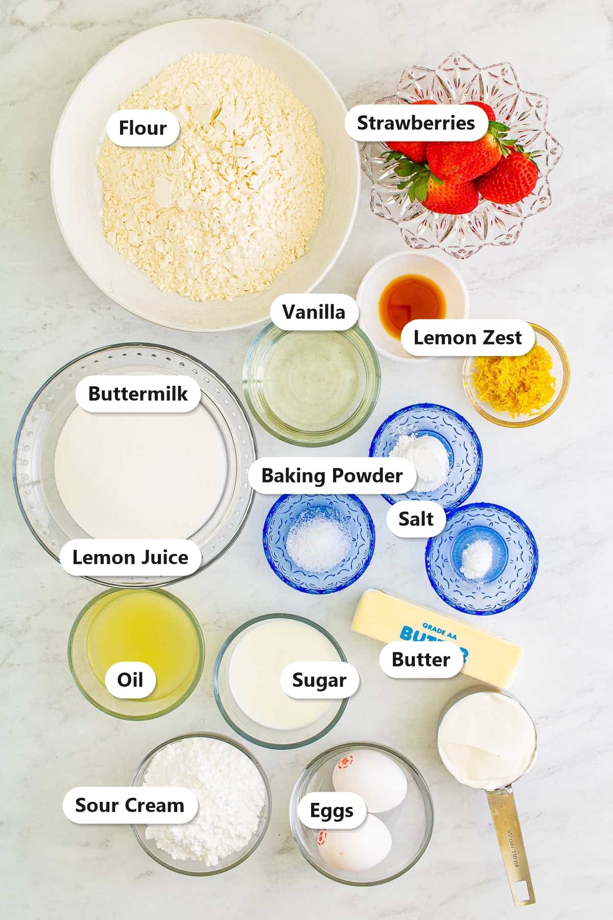 Labeled ingredients for lemon cupcakes with strawberry buttercream including flour, lemon, strawberries, butter, sugar, and eggs.