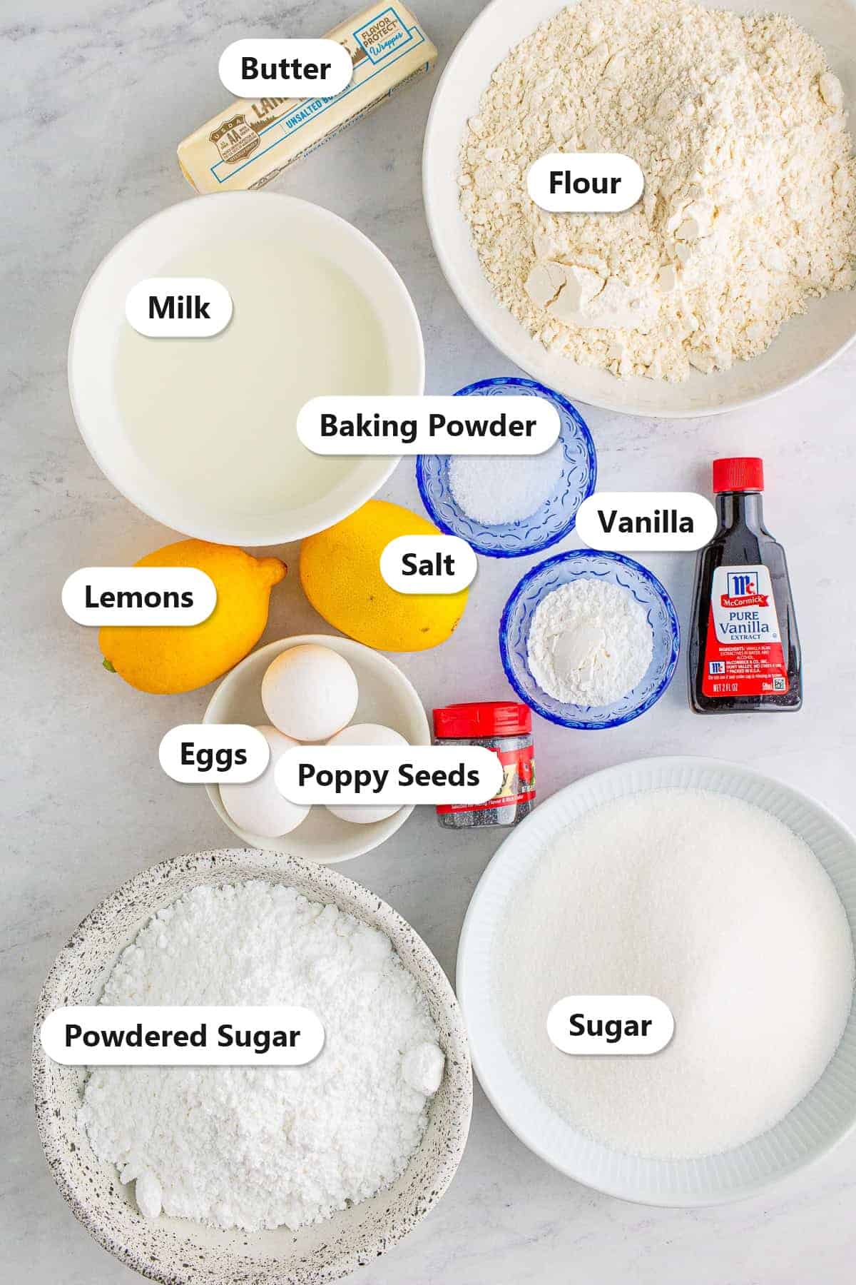 Labeled ingredients for Lemon Poppy Seed Cake including flour, butter, sugar, eggs, milk, lemons, poppy seeds, vanilla, and powdered sugar.
