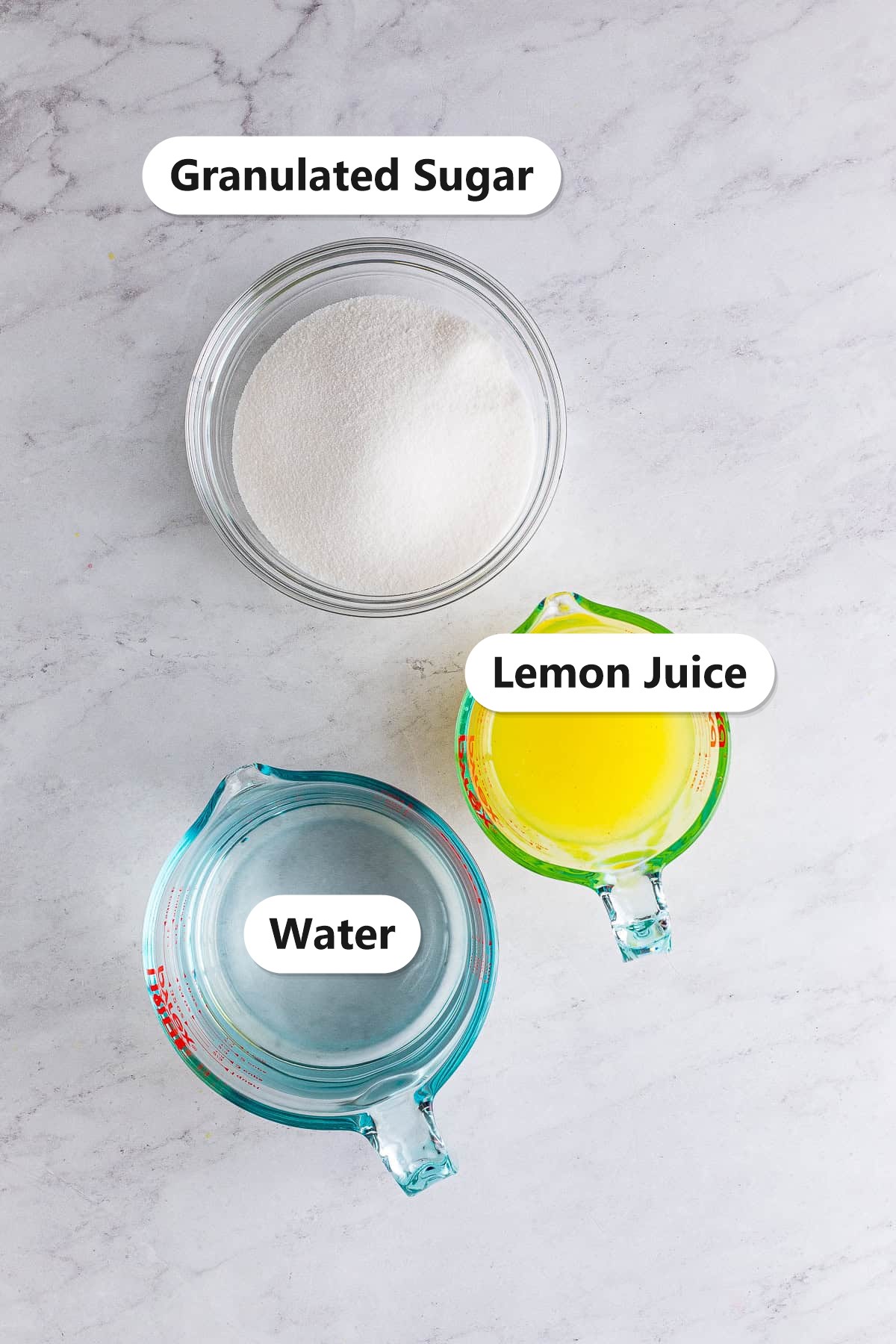 Ingredients to make homemade lemonade labeled on a marble surface: granulated sugar, fresh lemon juice, and water.