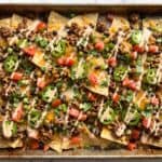 Loaded nachos on a sheet pan with melted cheese, ground beef, beans, jalapeno, tomato, and cilantro.