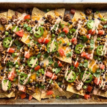Loaded nachos on a sheet pan with melted cheese, ground beef, beans, jalapeno, tomato, and cilantro.