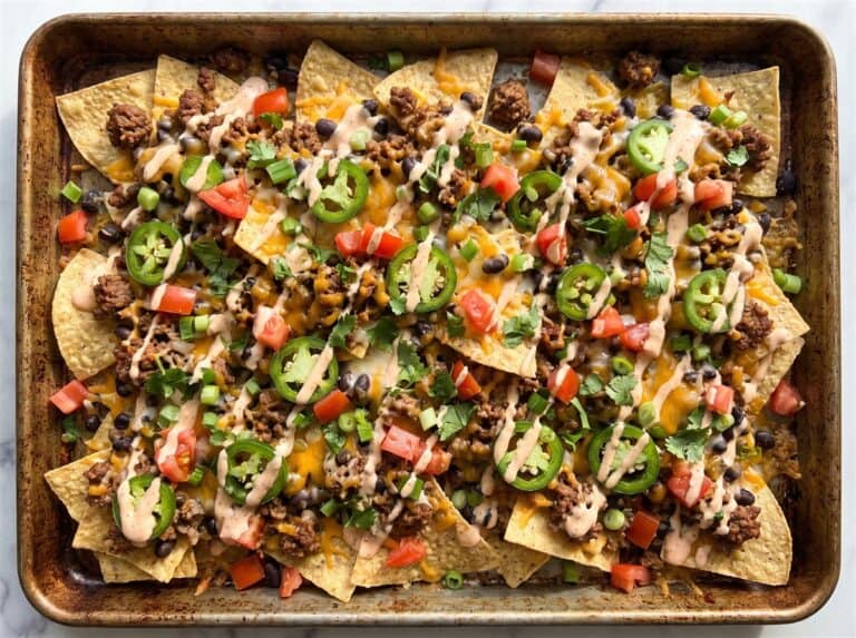 Loaded nachos on a sheet pan with melted cheese, ground beef, beans, jalapeno, tomato, and cilantro.