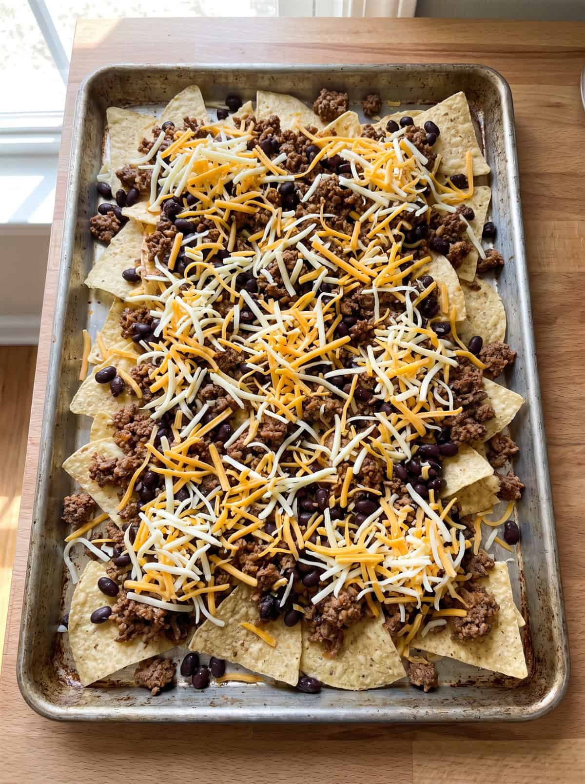 Tortilla chips layered with beef, beans, and cheese on a sheet pan before baking.