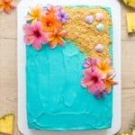 A luau cake frosted with turquoise ocean buttercream, graham cracker sand, and tropical flowers.