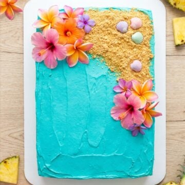 A luau cake frosted with turquoise ocean buttercream, graham cracker sand, and tropical flowers.