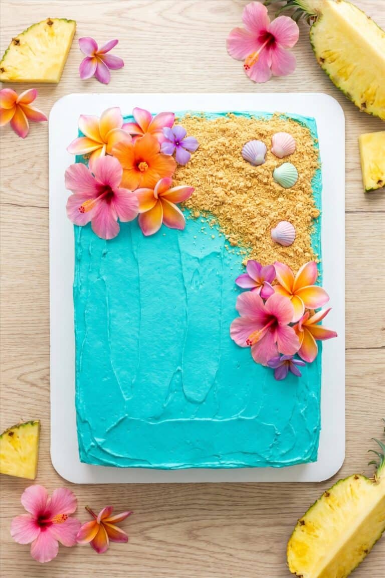 A luau cake frosted with turquoise ocean buttercream, graham cracker sand, and tropical flowers.