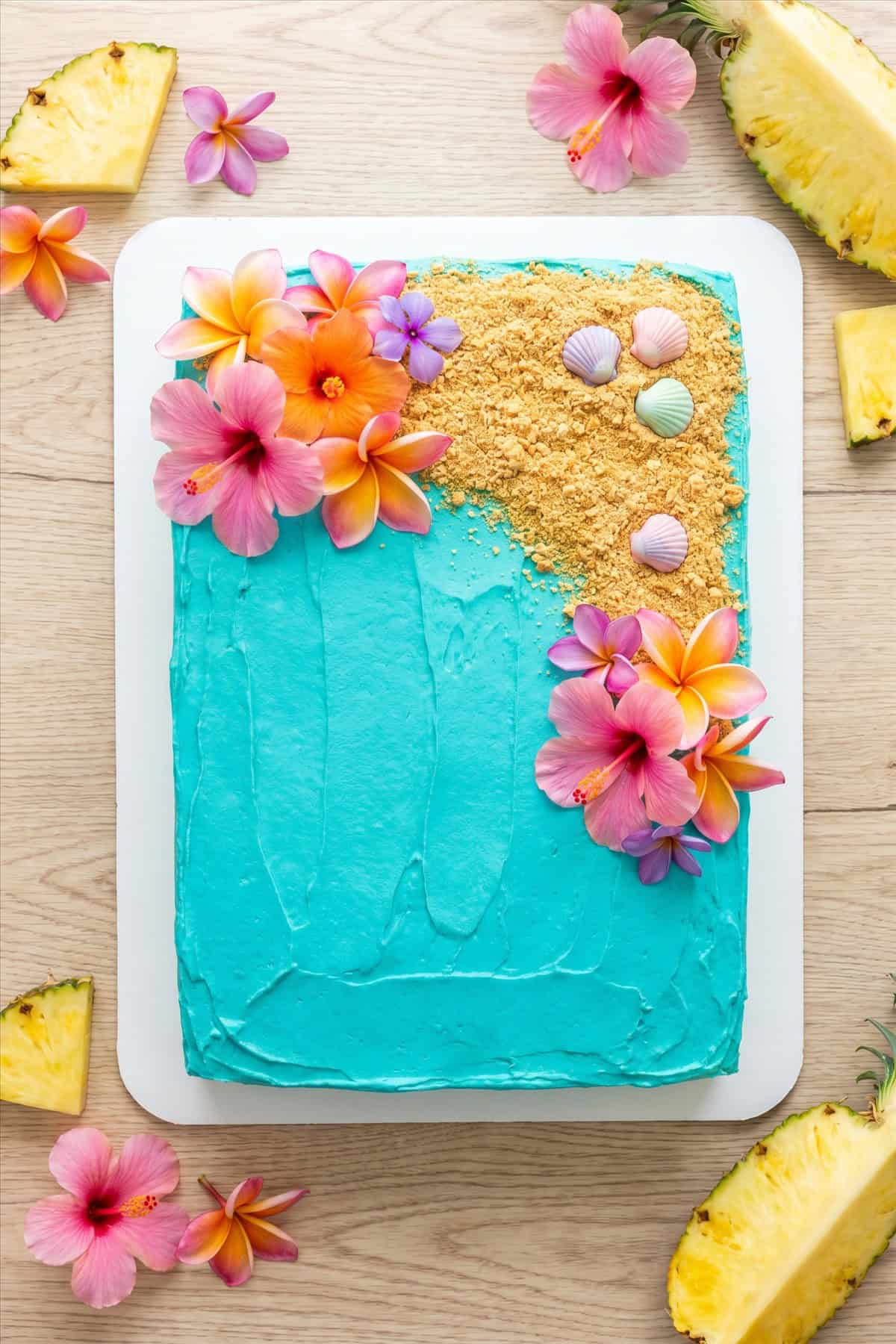 A luau cake frosted with turquoise ocean buttercream, graham cracker sand, and tropical flowers.