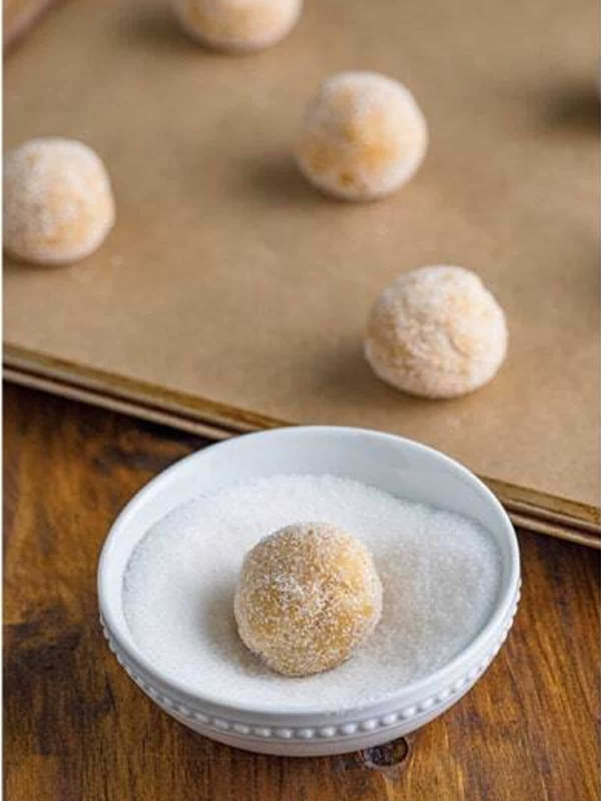 A chilled maple cookie dough ball being rolled in a small bowl of granulated sugar.