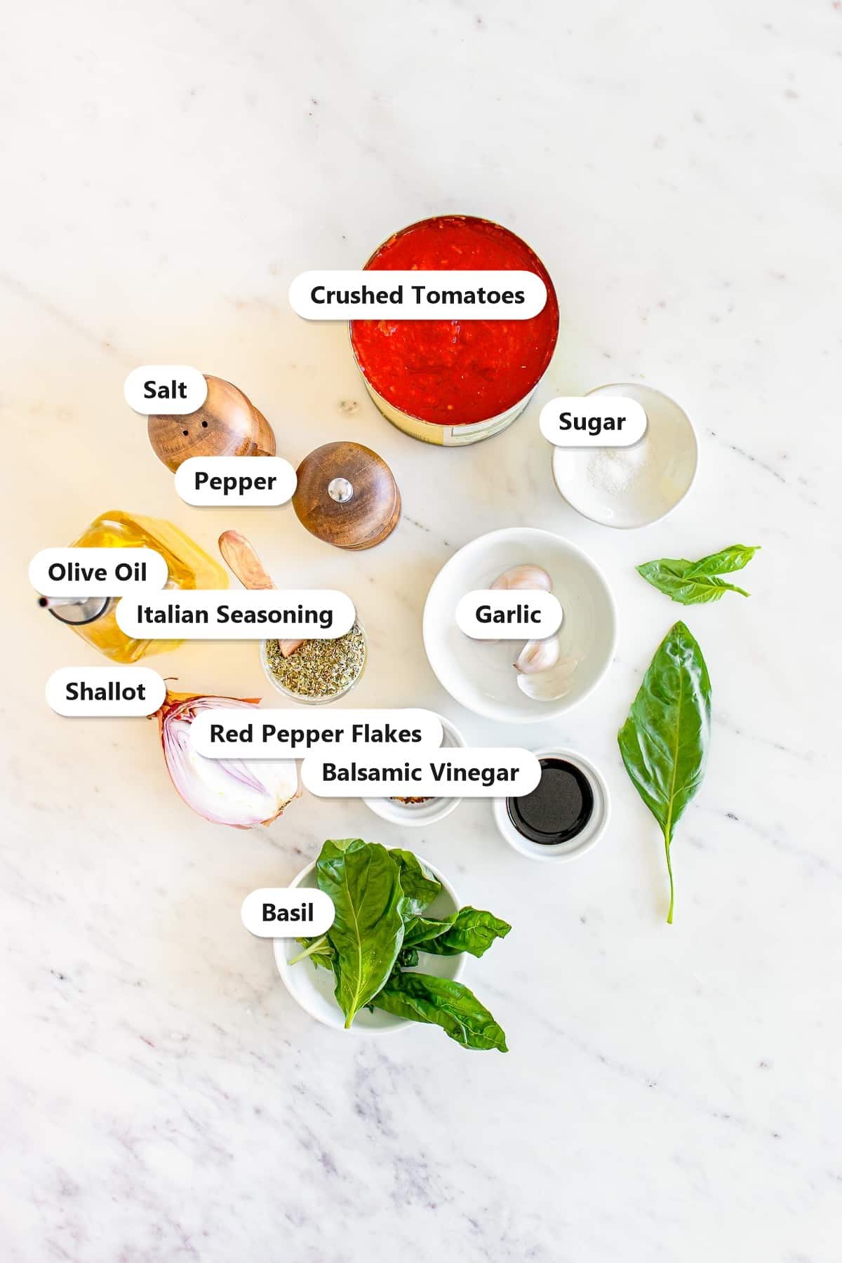 Labeled ingredients to make marinara sauce including crushed tomatoes, olive oil, shallot, garlic, salt, pepper, sugar, balsamic vinegar, Italian seasoning, red pepper flakes, and fresh basil.