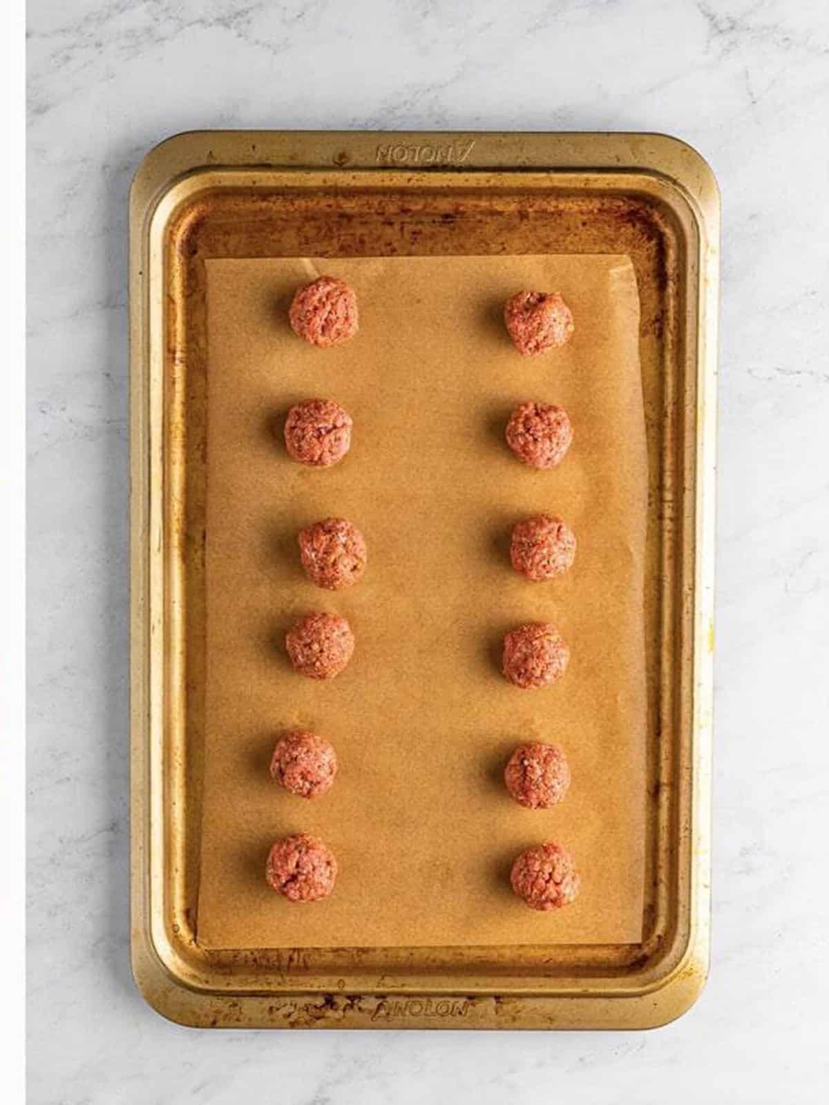 Seasoned ground beef rolled into meatballs on a lined baking sheet.