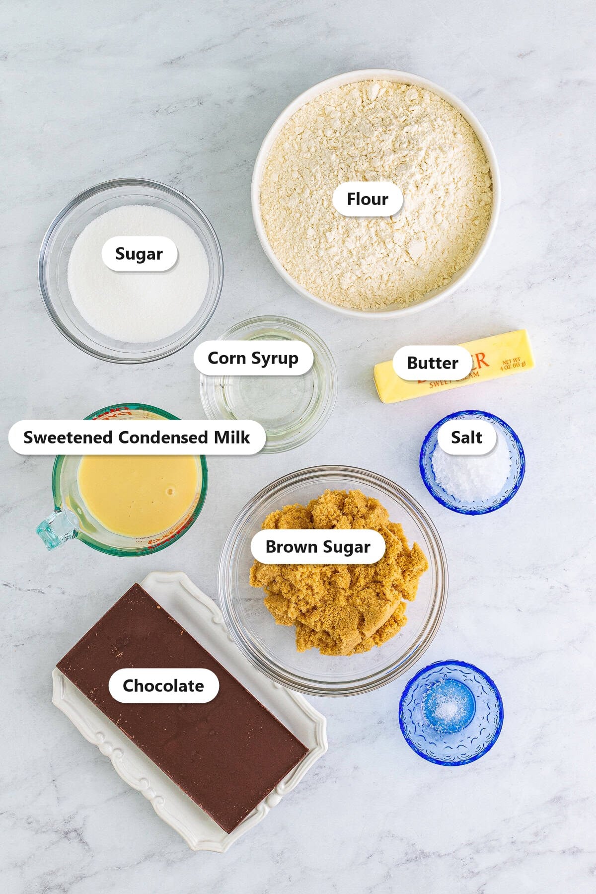 Labeled ingredients for millionaires shortbread including flour, butter, sugar, brown sugar, sweetened condensed milk, and chocolate.