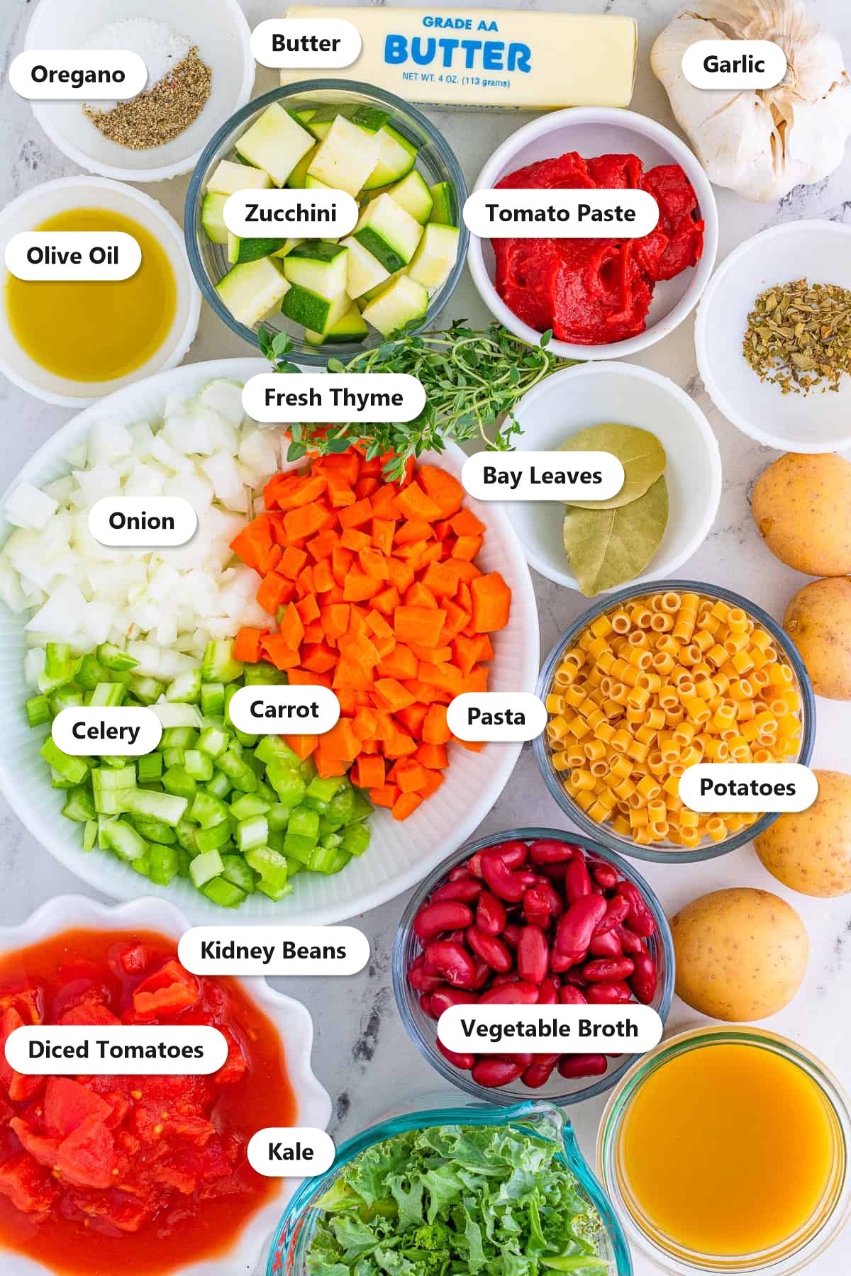 Minestrone soup ingredients including onion, carrot, celery, zucchini, potatoes, kidney beans, kale, pasta, diced tomatoes, and vegetable broth labeled on a marble surface.