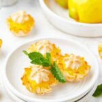 Mini lemon meringue pies on a white plate garnished with fresh mint and lemon slices.