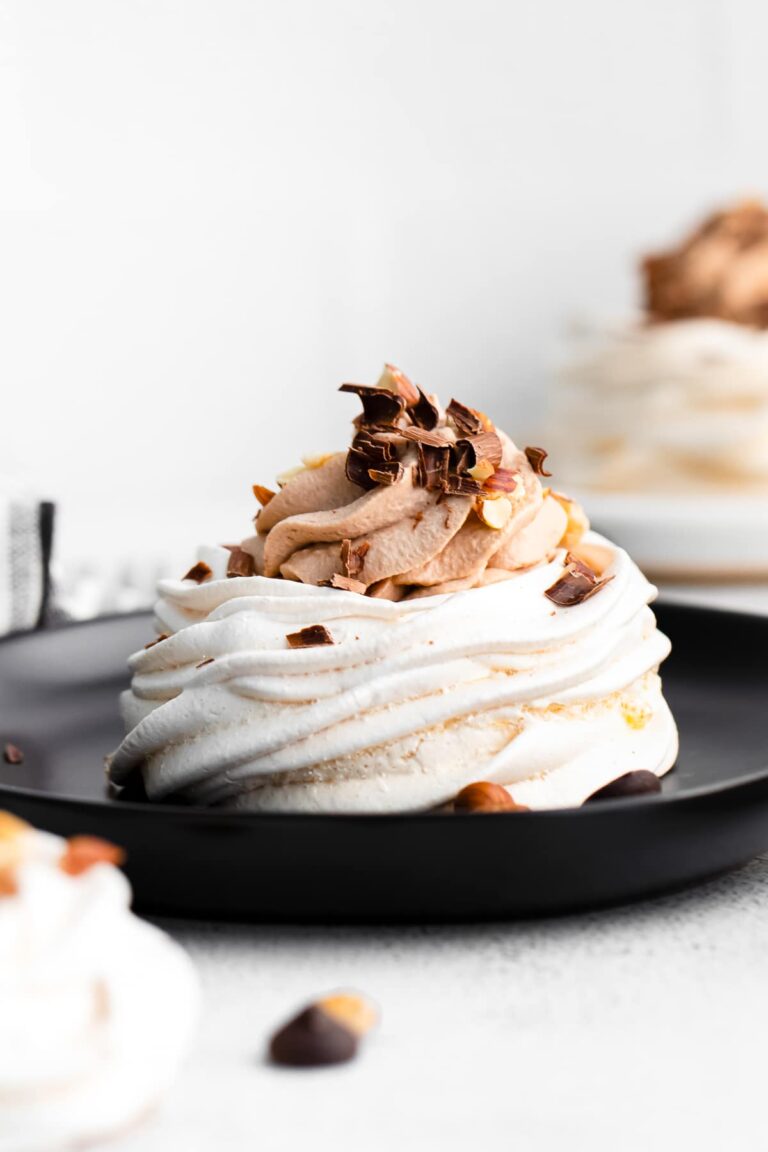 A mini pavlova topped with Nutella whipped cream and chopped hazelnuts on a black plate.