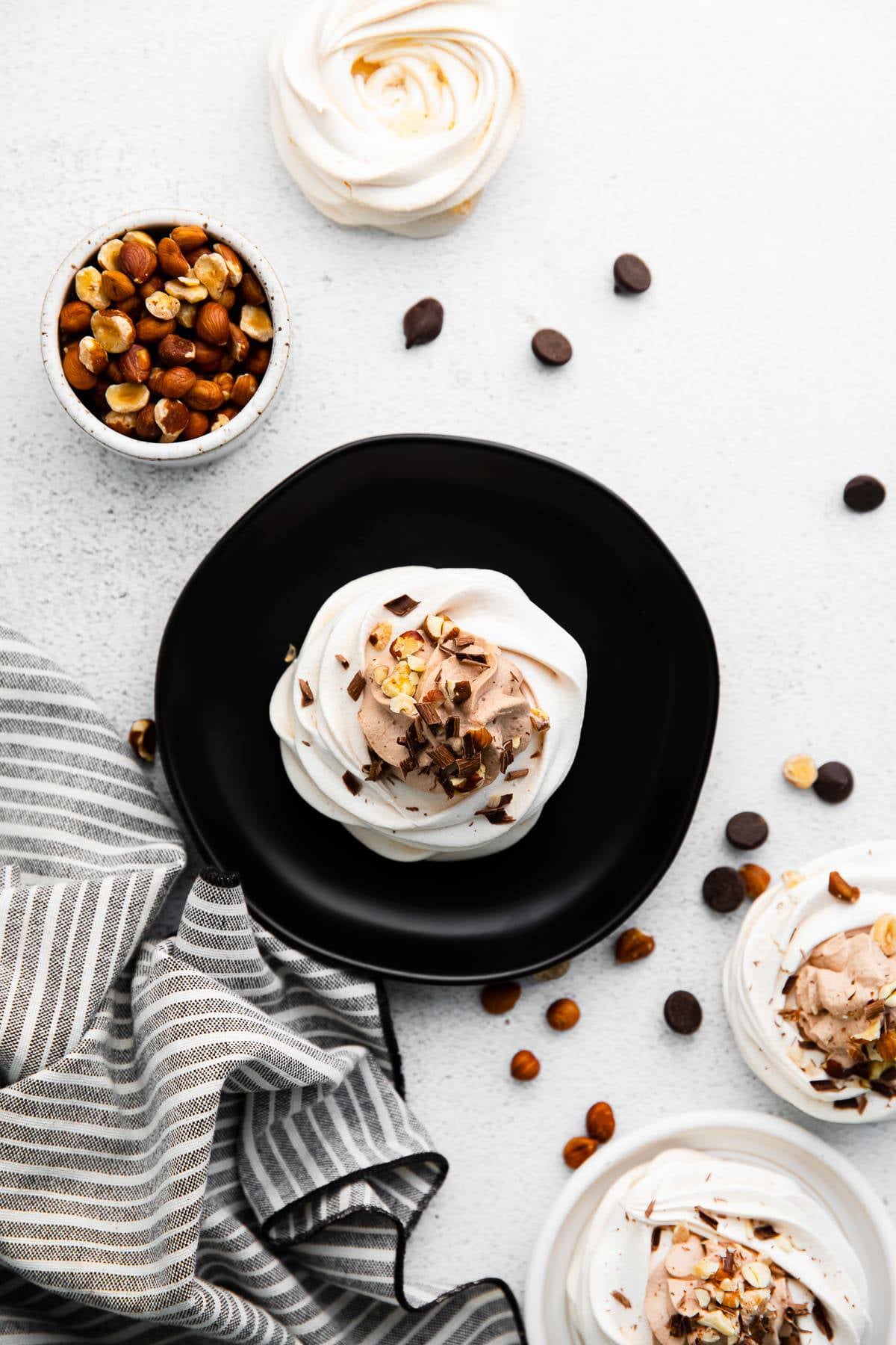 A mini pavlova on a black plate served alongside a bowl of toasted hazelnuts.