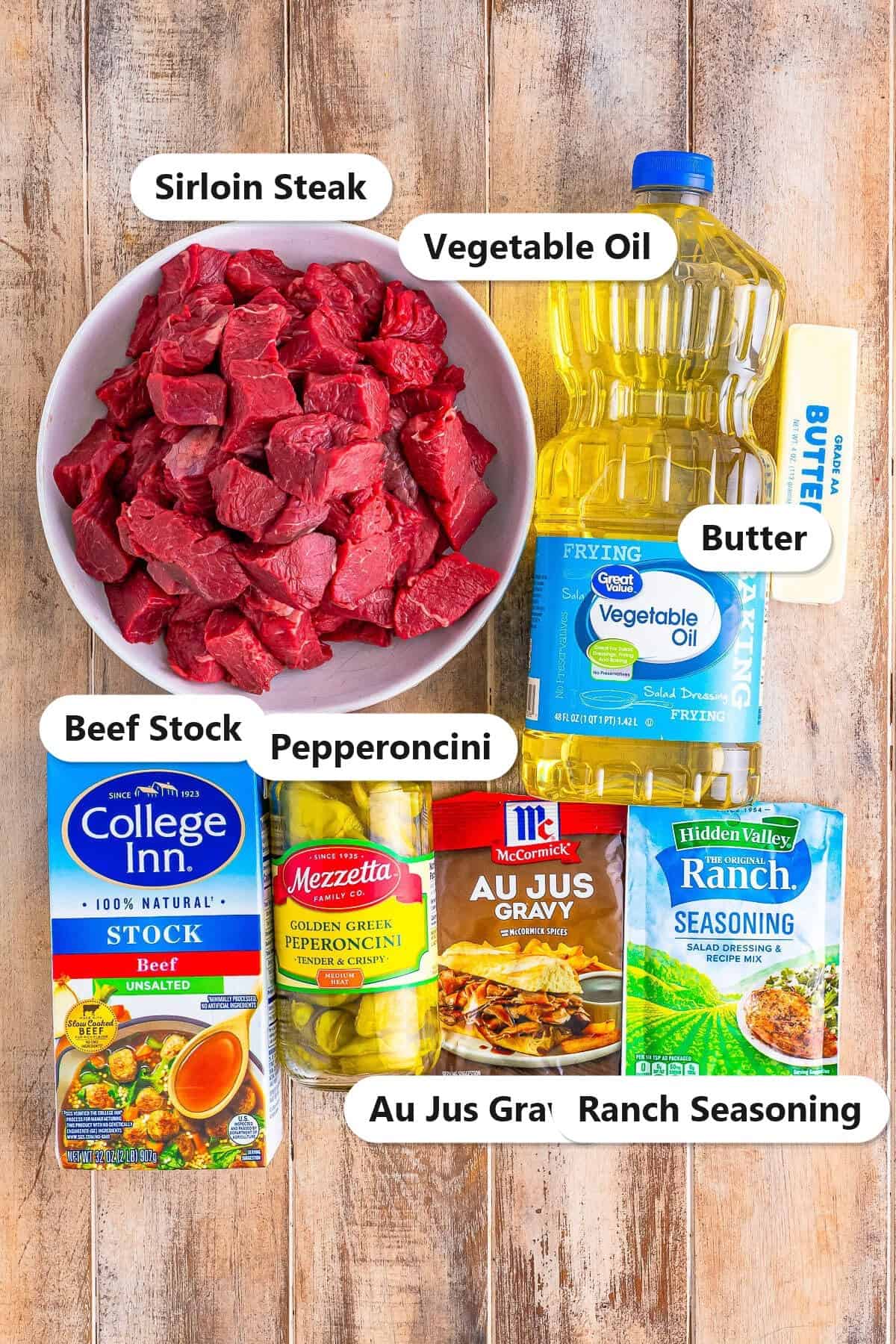 Labeled ingredients for slow cooker Mississippi steak bites including sirloin steak, vegetable oil, butter, beef stock, pepperoncini, au jus gravy mix, and ranch seasoning.
