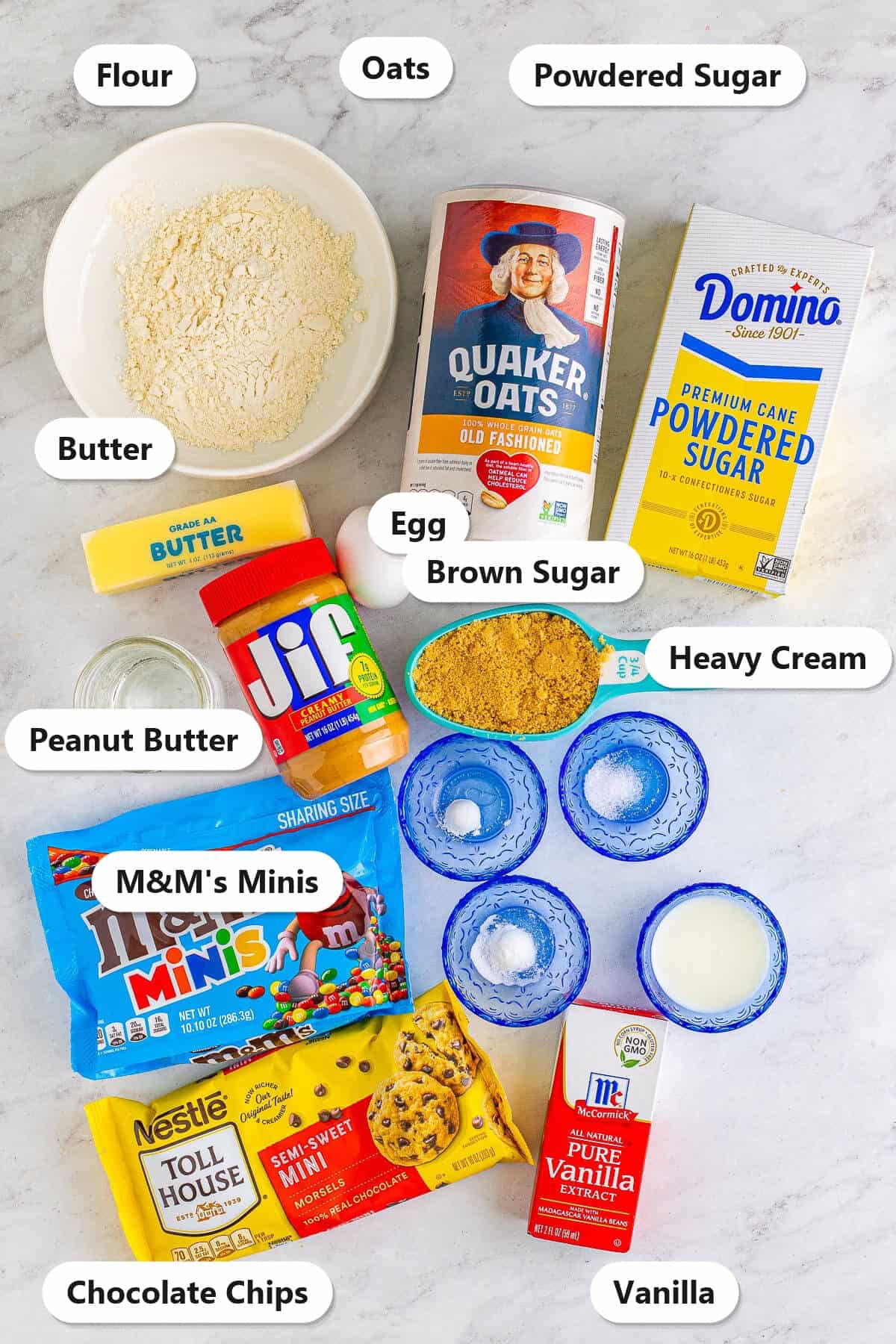 Labeled ingredients for monster cookie cupcakes including oats, peanut butter, flour, brown sugar, M&M's minis, chocolate chips, powdered sugar, and vanilla.