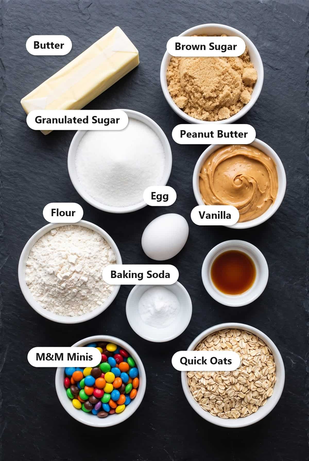 Labeled ingredients for monster cookies including butter, brown sugar, granulated sugar, peanut butter, egg, flour, vanilla, baking soda, mini candy-coated chocolates, and quick oats.