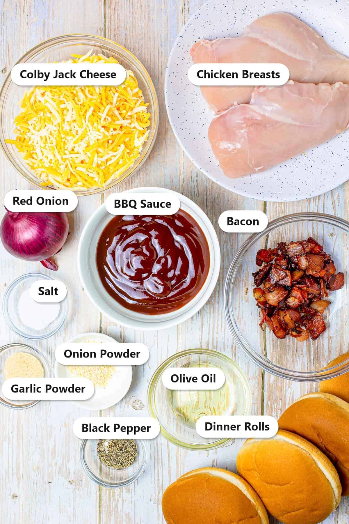 Ingredients to make monterey chicken sliders labeled including chicken breasts, colby jack cheese, BBQ sauce, red onion, bacon, dinner rolls, olive oil, and seasonings.
