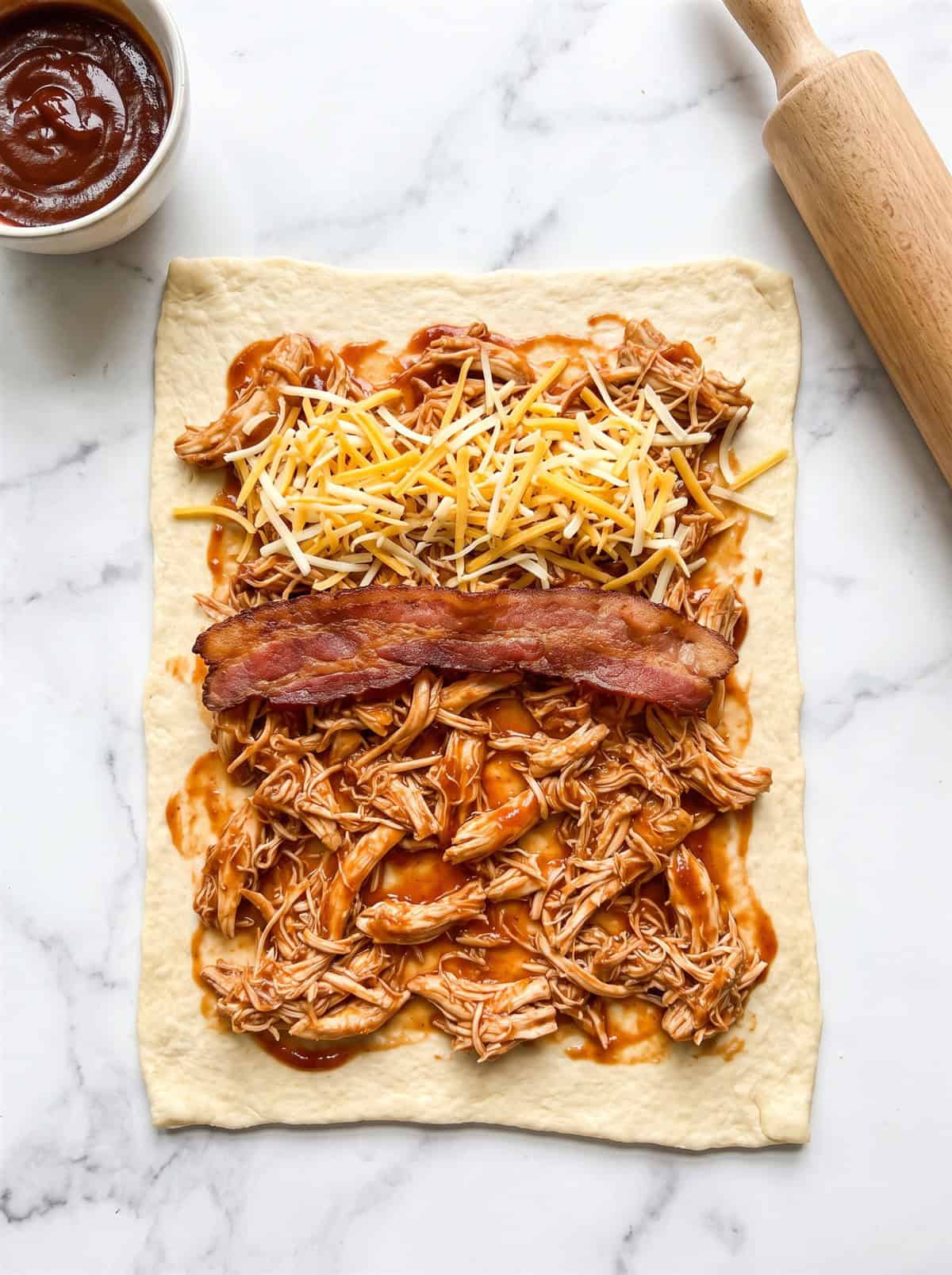 Crescent roll dough rectangle topped with BBQ shredded chicken, bacon, and shredded cheese ready to roll up.