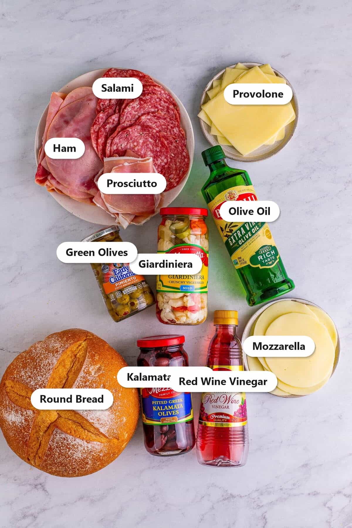 Labeled ingredients to make a muffuletta: salami, ham, prosciutto, provolone, olive oil, giardiniera, green olives, mozzarella, round bread, kalamata olives, and red wine vinegar.