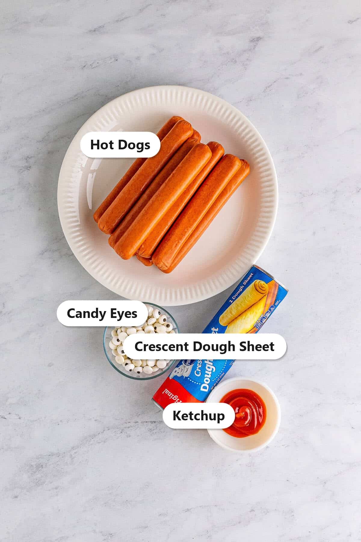 Labeled ingredients for mummy dogs including hot dogs, crescent dough sheet, candy eyes, and ketchup.