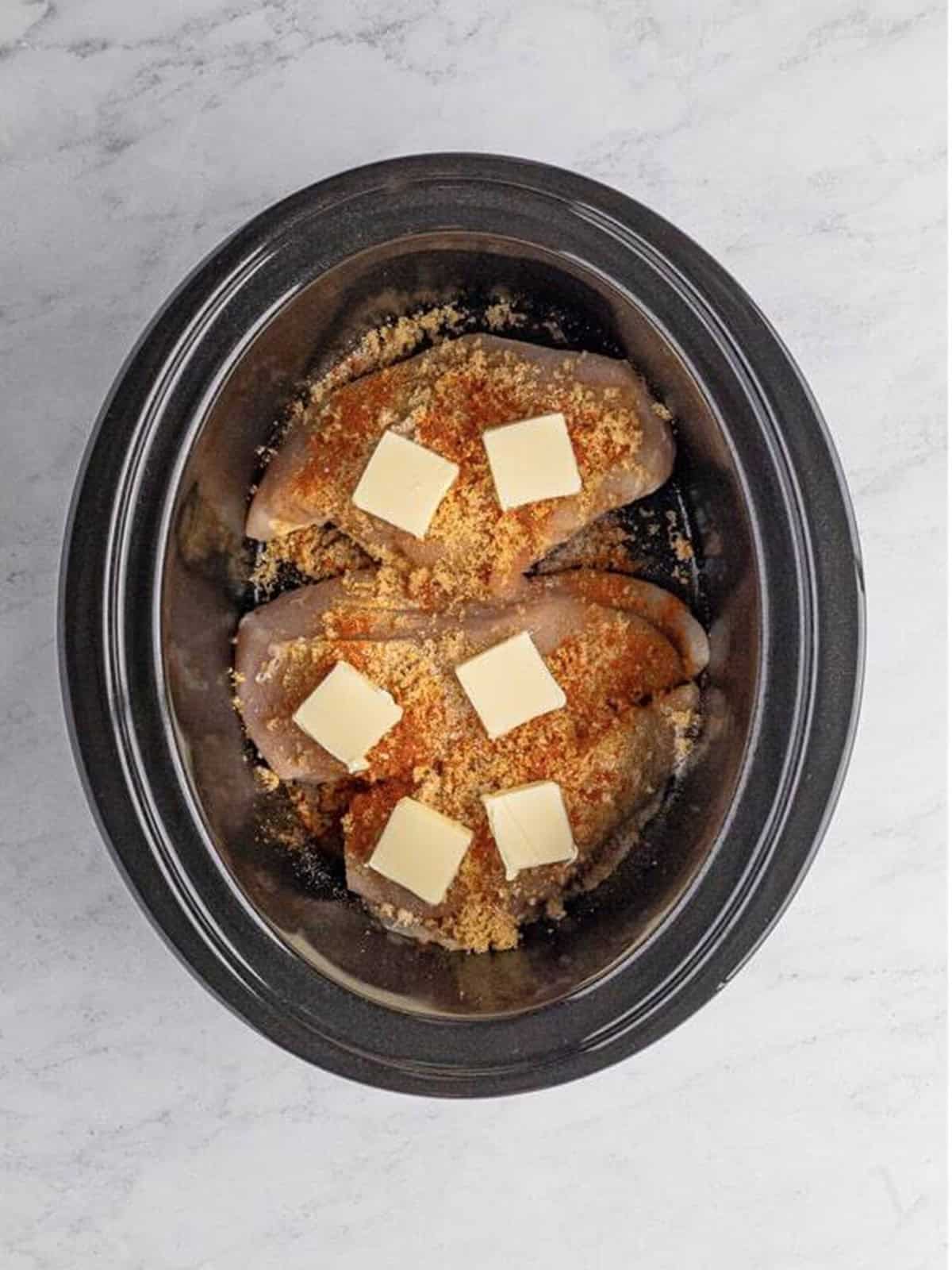 Seasoned chicken in a slow cooker topped with slices of butter.