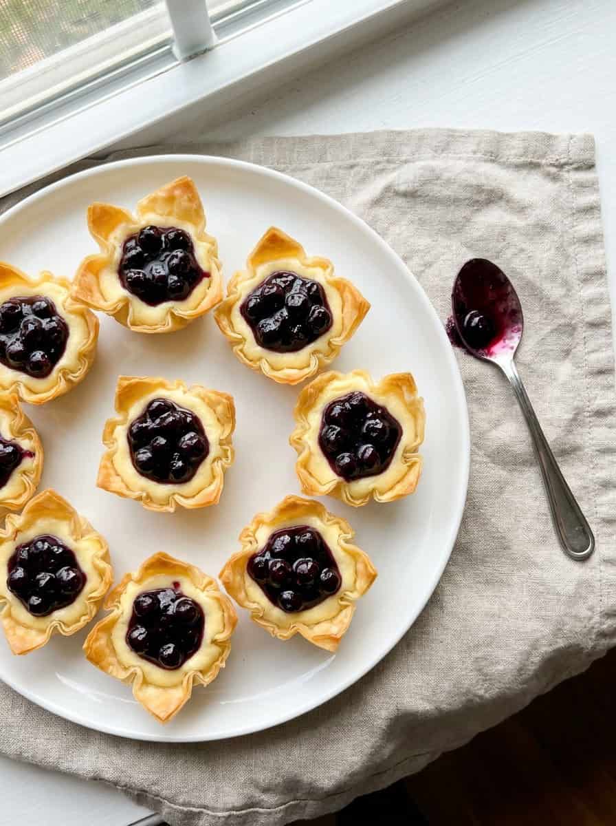 Cheesecake-filled phyllo cups being topped with spoonfuls of wild blueberry sauce.