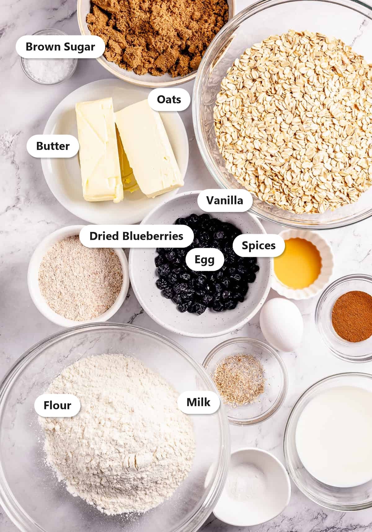Labeled ingredients for oatmeal blueberry cookies including brown sugar, oats, butter, dried blueberries, vanilla, egg, spices, flour, and milk.