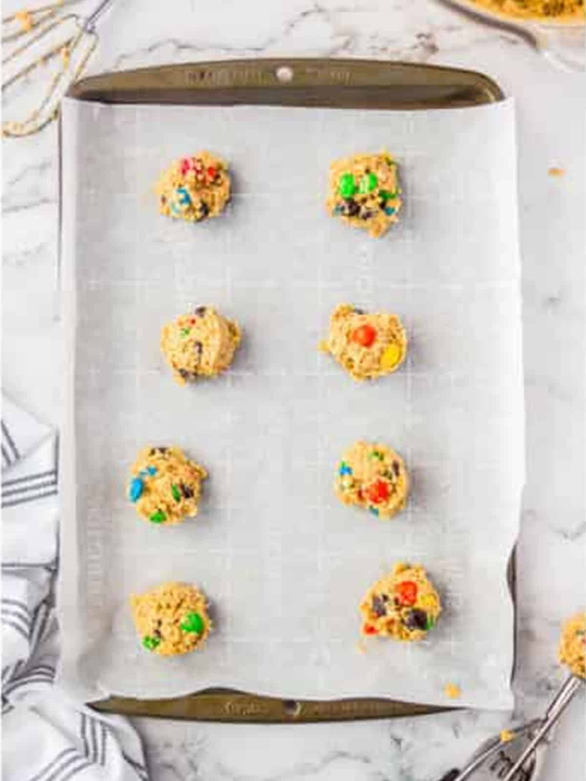 Rounded scoops of oatmeal M&M cookie dough spaced on a parchment-lined sheet tray.