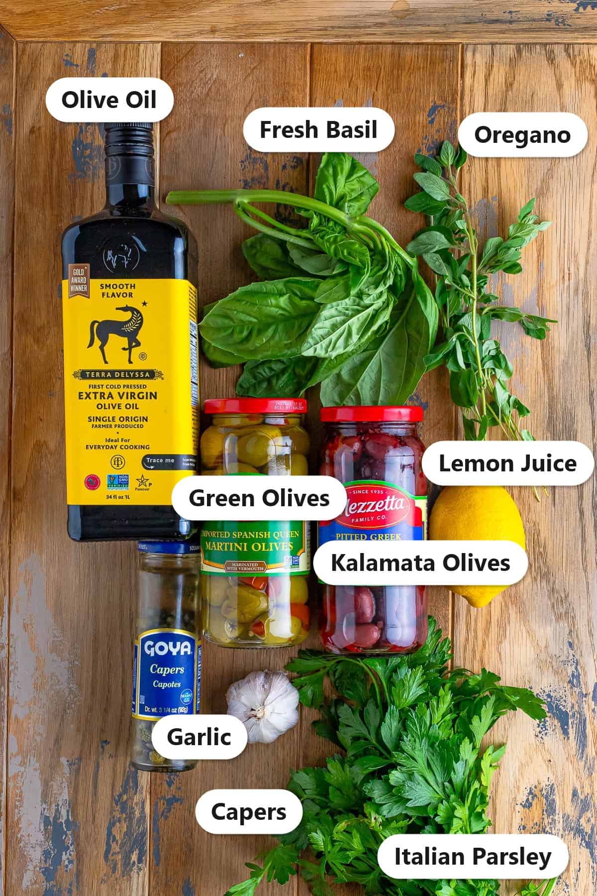 Labeled ingredients for olive tapenade recipe including green olives, kalamata olives, capers, olive oil, garlic, lemon juice, fresh basil, oregano, and Italian parsley.