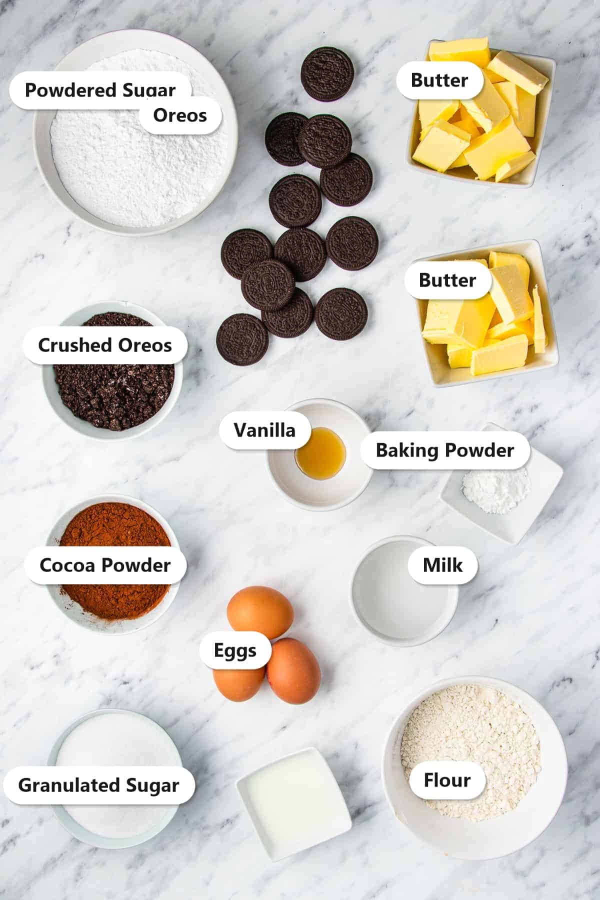 Labeled ingredients to make Oreo cupcakes including cocoa powder, flour, butter, eggs, powdered sugar, milk, crushed Oreos, and whole Oreos.