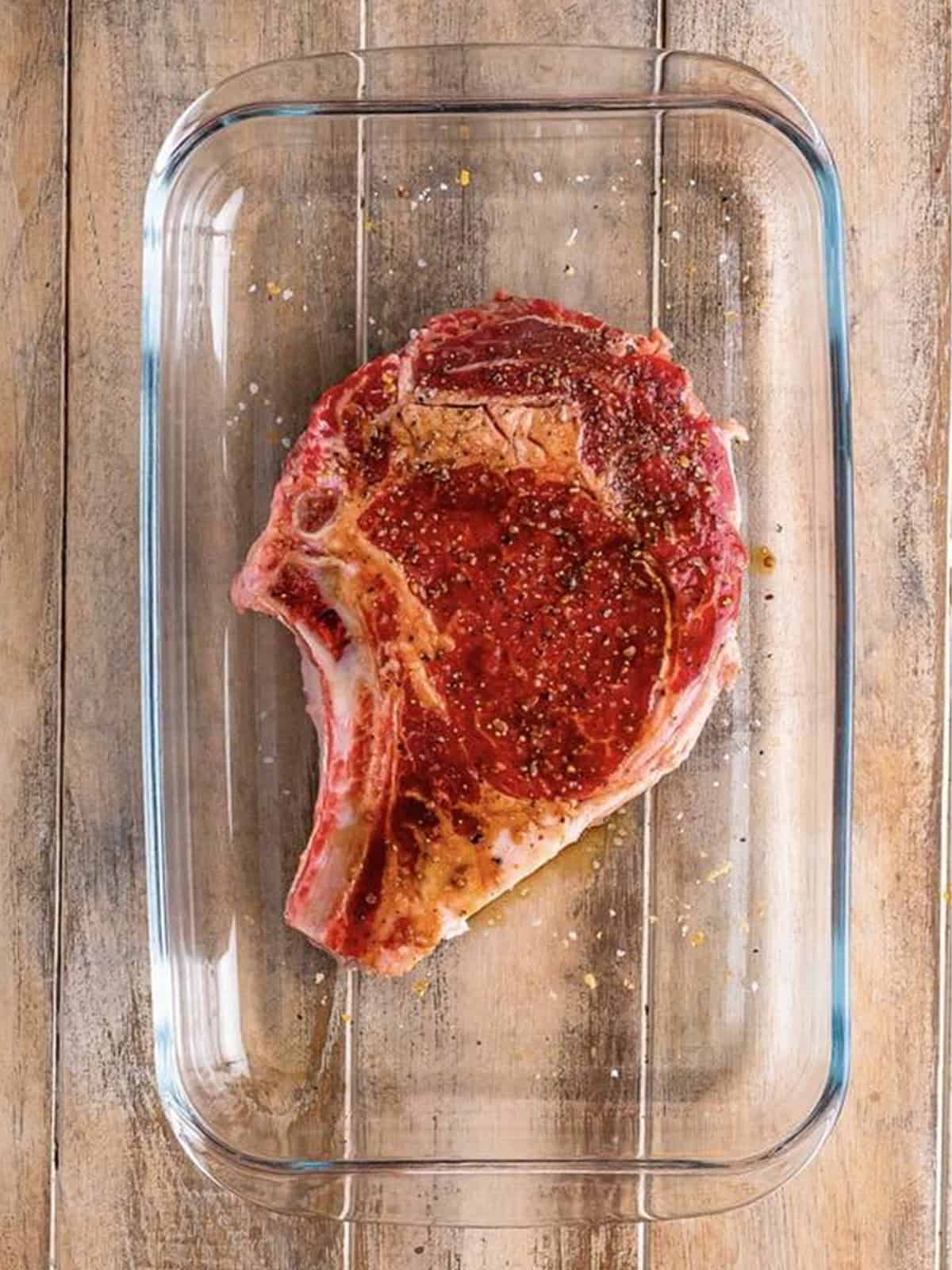 Bone-in ribeye steak seasoned with brown sugar and steak seasoning in a glass dish.