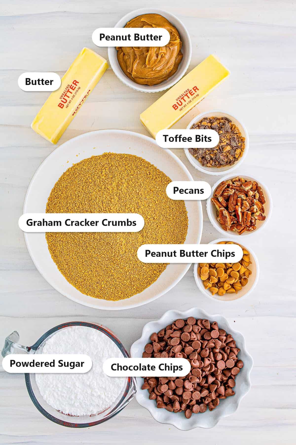 Labeled ingredients to make no-bake peanut butter bars: peanut butter, butter, toffee bits, pecans, peanut butter chips, graham cracker crumbs, powdered sugar, and chocolate chips.