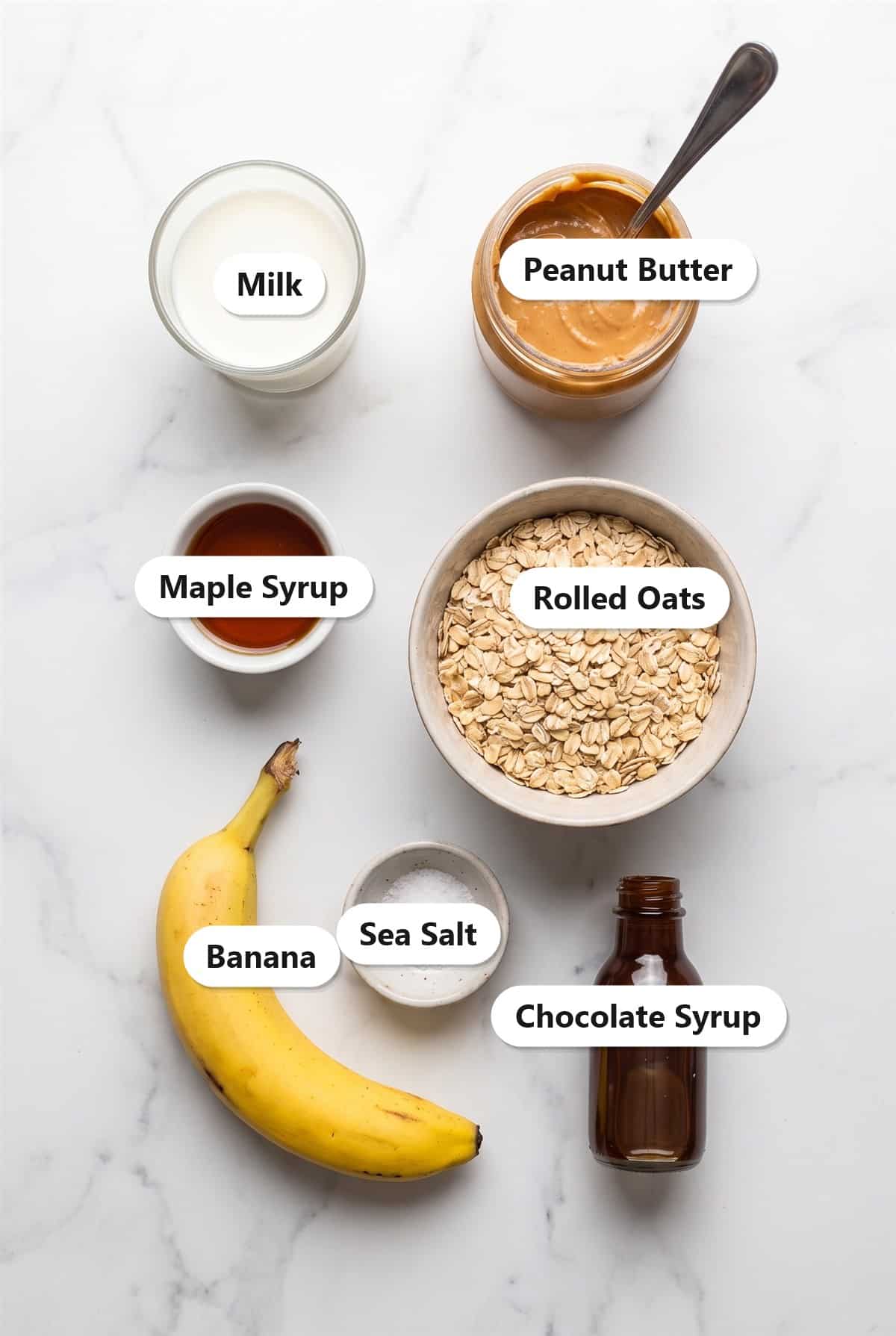 Labeled ingredients for peanut butter overnight oats including milk, peanut butter, maple syrup, rolled oats, banana, sea salt, and chocolate syrup.