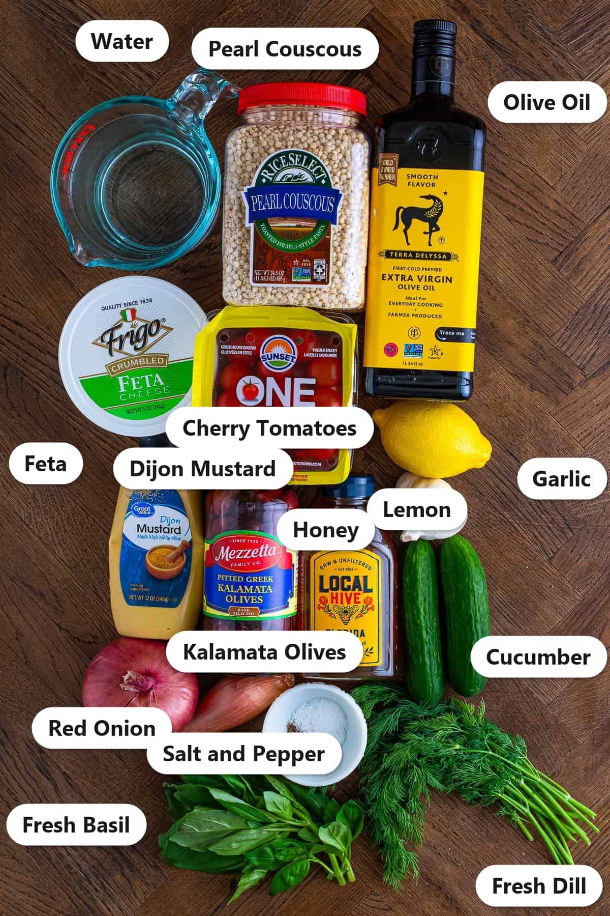 Labeled ingredients for pearl couscous salad including pearl couscous, olive oil, feta, cherry tomatoes, kalamata olives, cucumber, red onion, lemon, garlic, dijon mustard, honey, fresh dill, and basil.