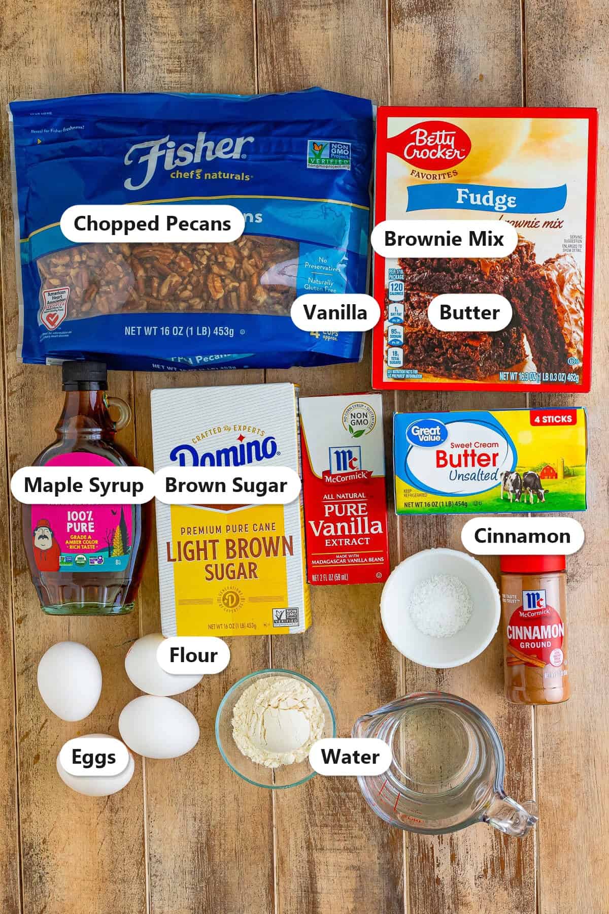 Labeled ingredients for chocolate pecan cobbler including chopped pecans, brownie mix, maple syrup, brown sugar, butter, eggs, vanilla, cinnamon, flour, and water.