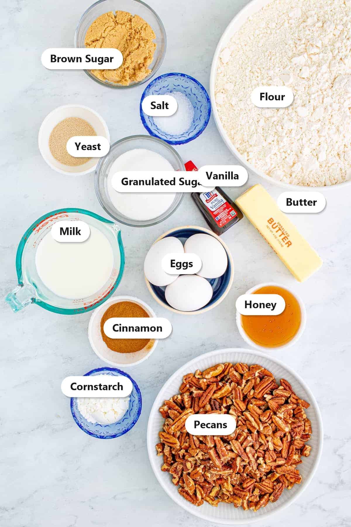 Labeled ingredients for pecan sticky buns including flour, yeast, milk, butter, eggs, brown sugar, cinnamon, honey, and pecans.