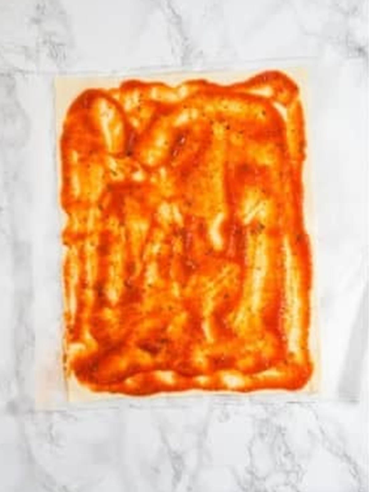 Marinara sauce spread over a sheet of puff pastry leaving a border around the edges.