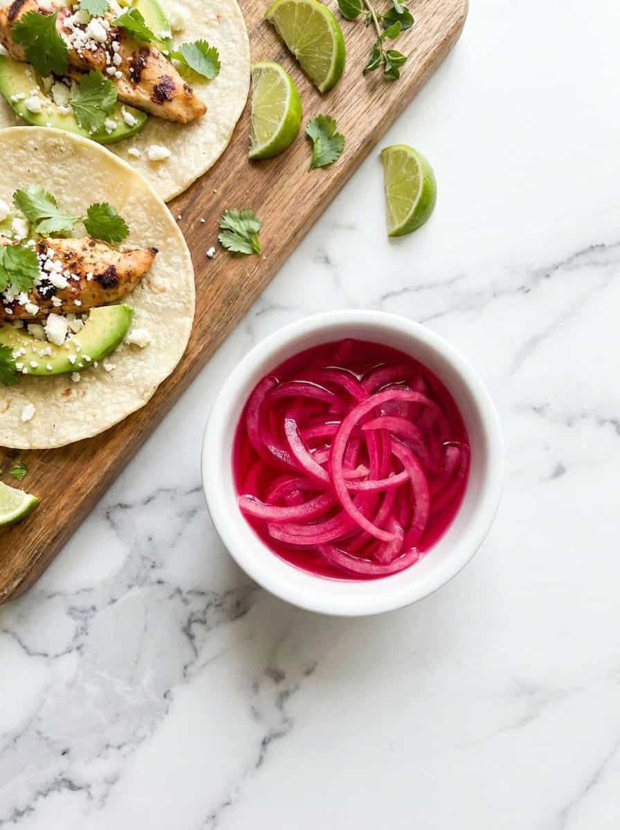 A small bowl of pickled red onions next to fresh tacos.