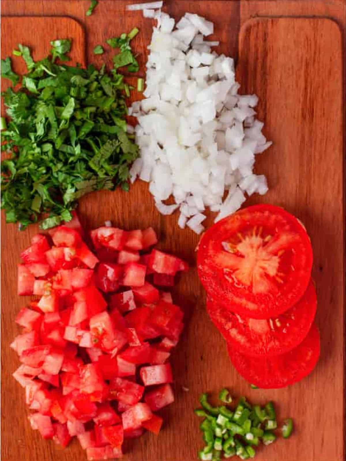 Finely chopped Roma tomatoes, onion, cilantro, and serrano chilis on a wooden cutting board.